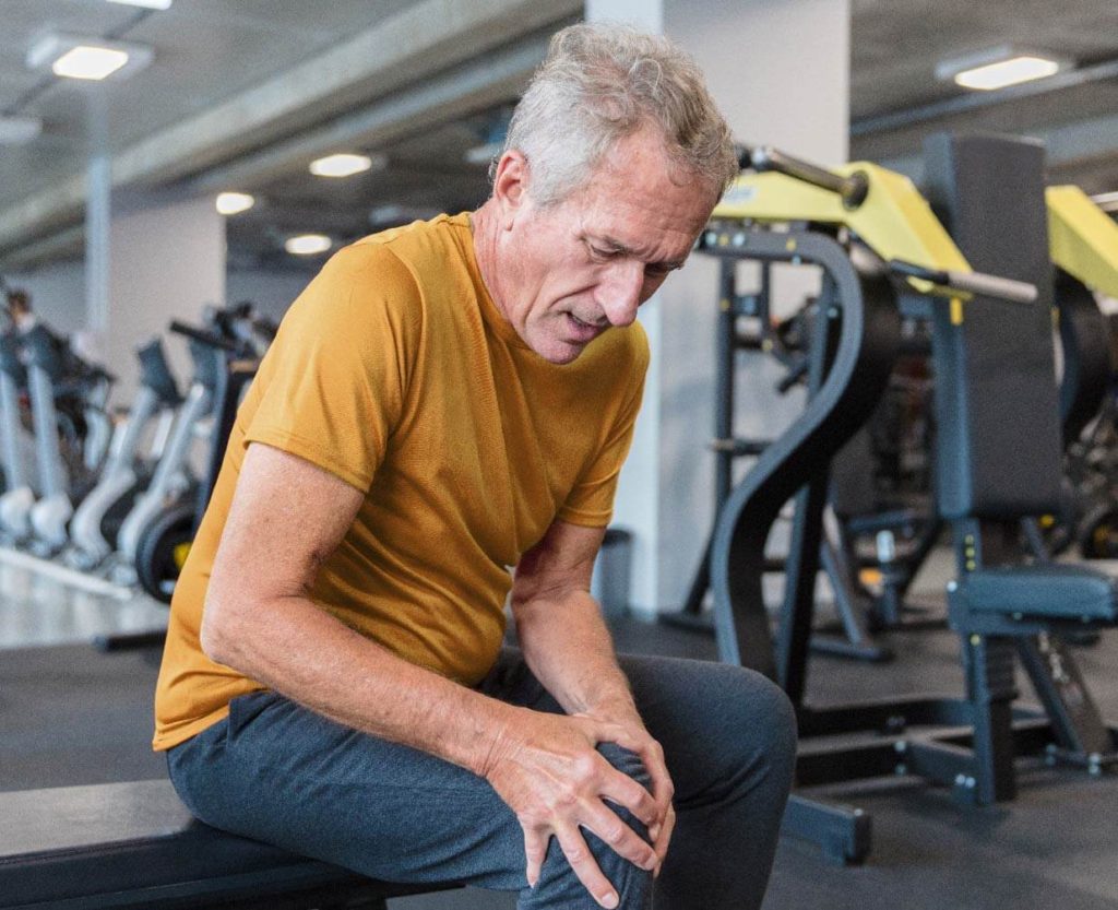Man in a gym, taking a break sitting on a bench holding his knee which appears to be in pain